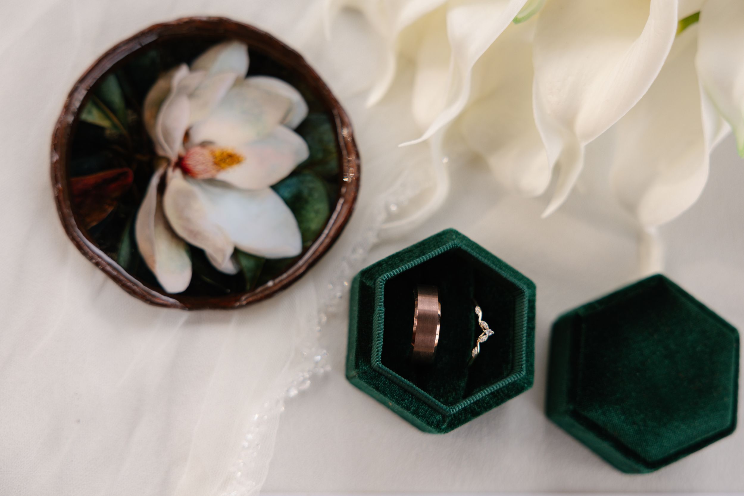 Wedding rings in dark green velvet hexagonal box next to white magnolia flower on light surface.