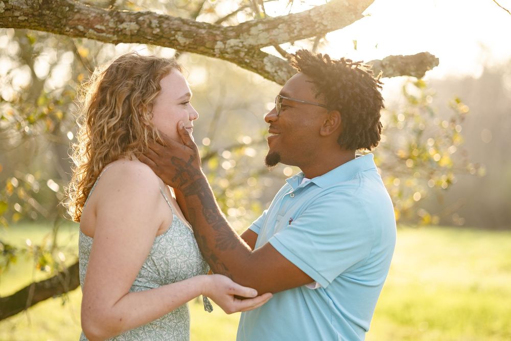 A couple in formal attire dances and laughs together in a sunlit outdoor setting with trees in the background.