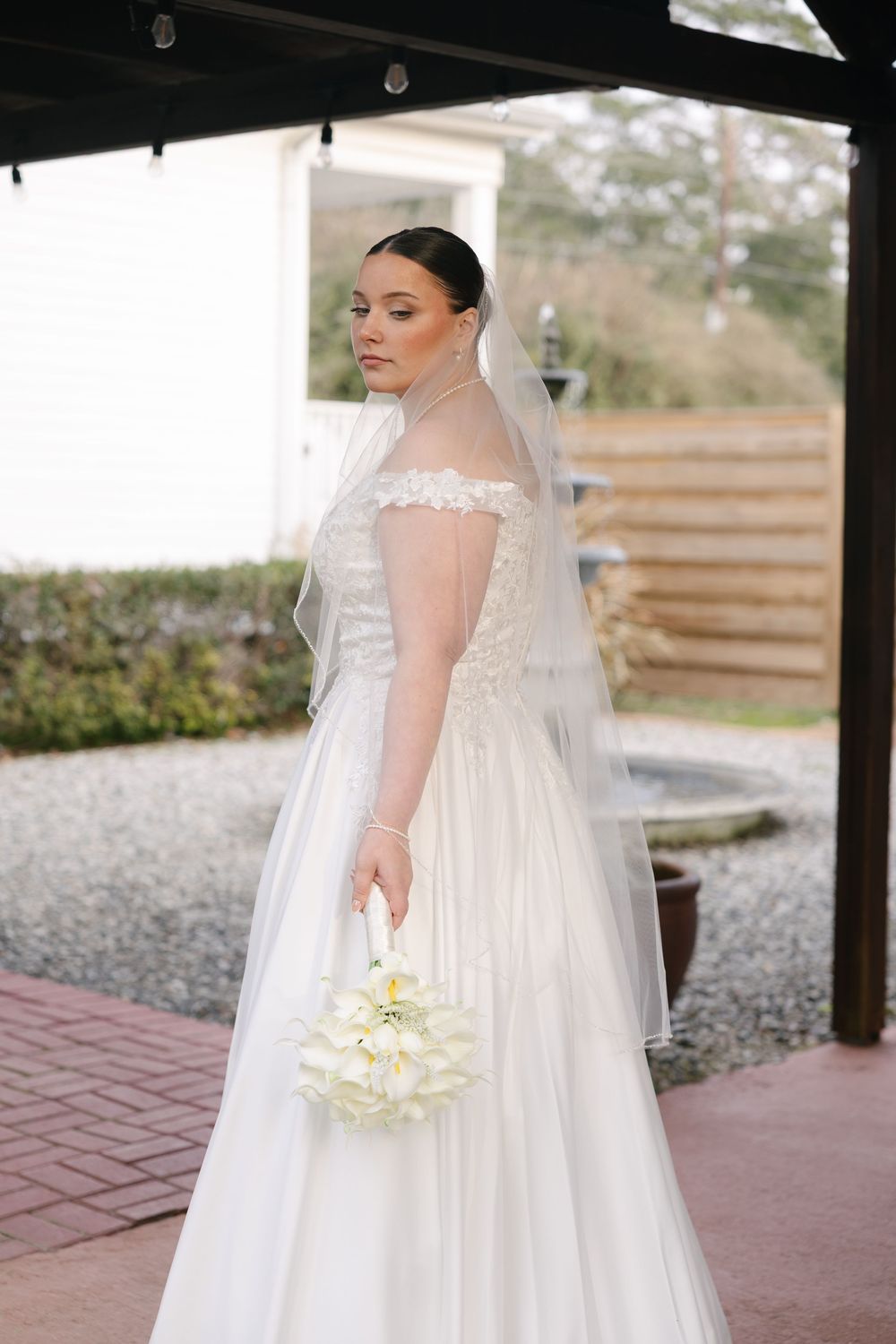 A bride in an elegant white empire waist wedding gown with flowing veil holds a white bouquet during an outdoor photoshoot.