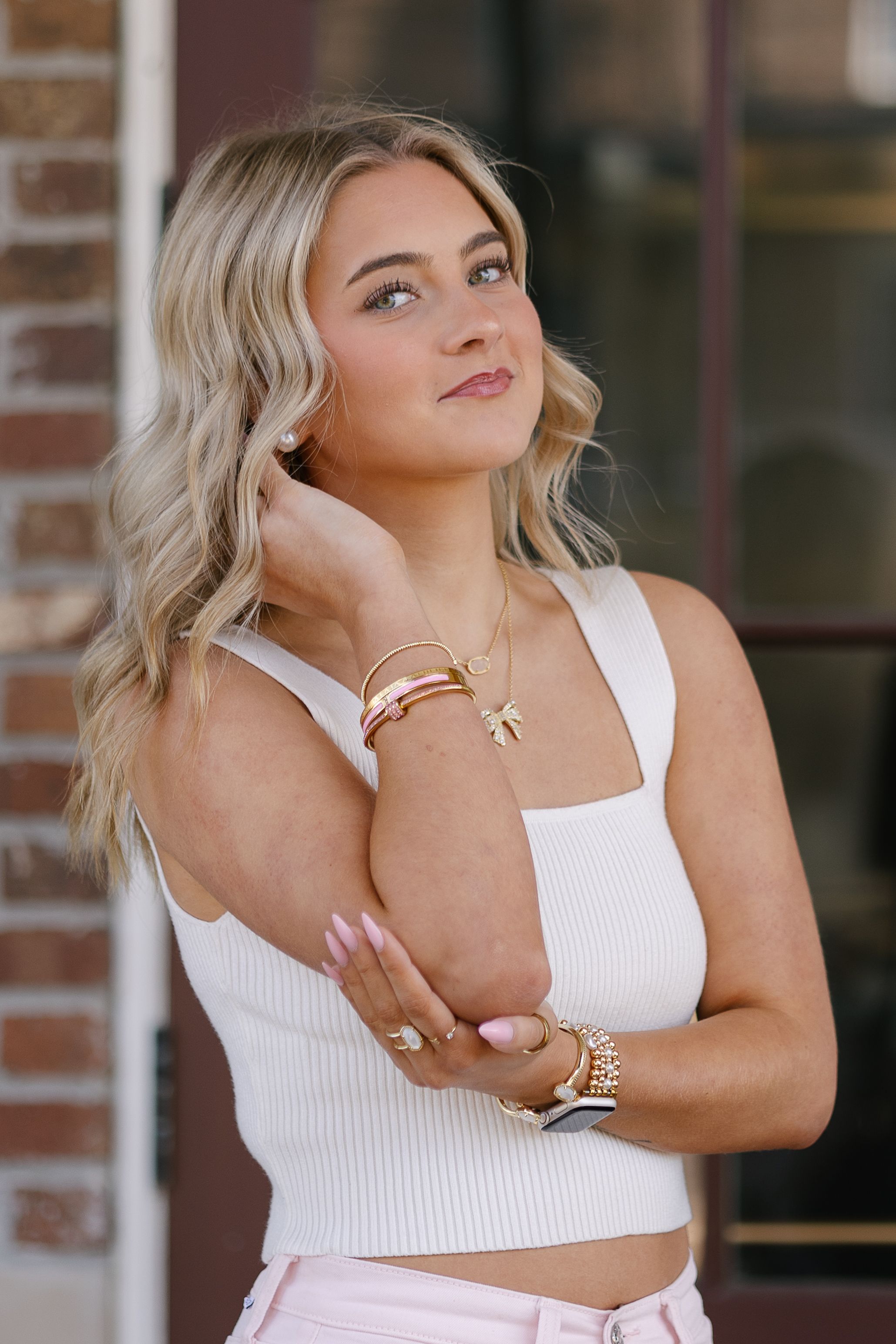 Woman in white tank top poses against brick wall wearing gold jewelry and bracelets.