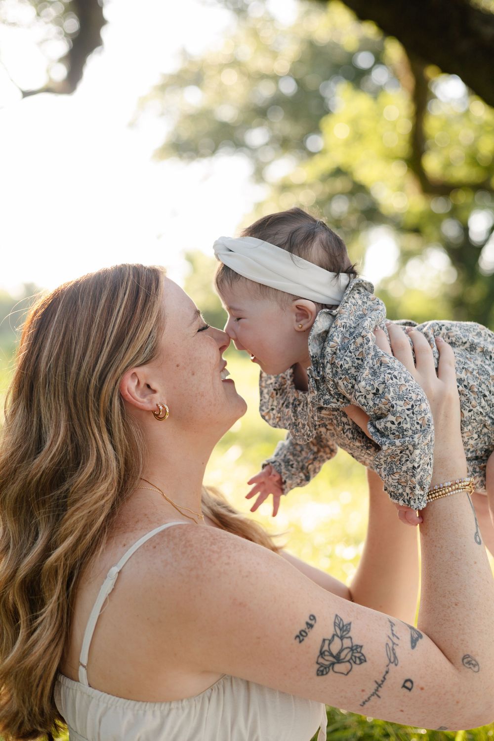 A tender moment captured outdoors as a parent lifts up their baby wearing a floral outfit in golden sunlight.