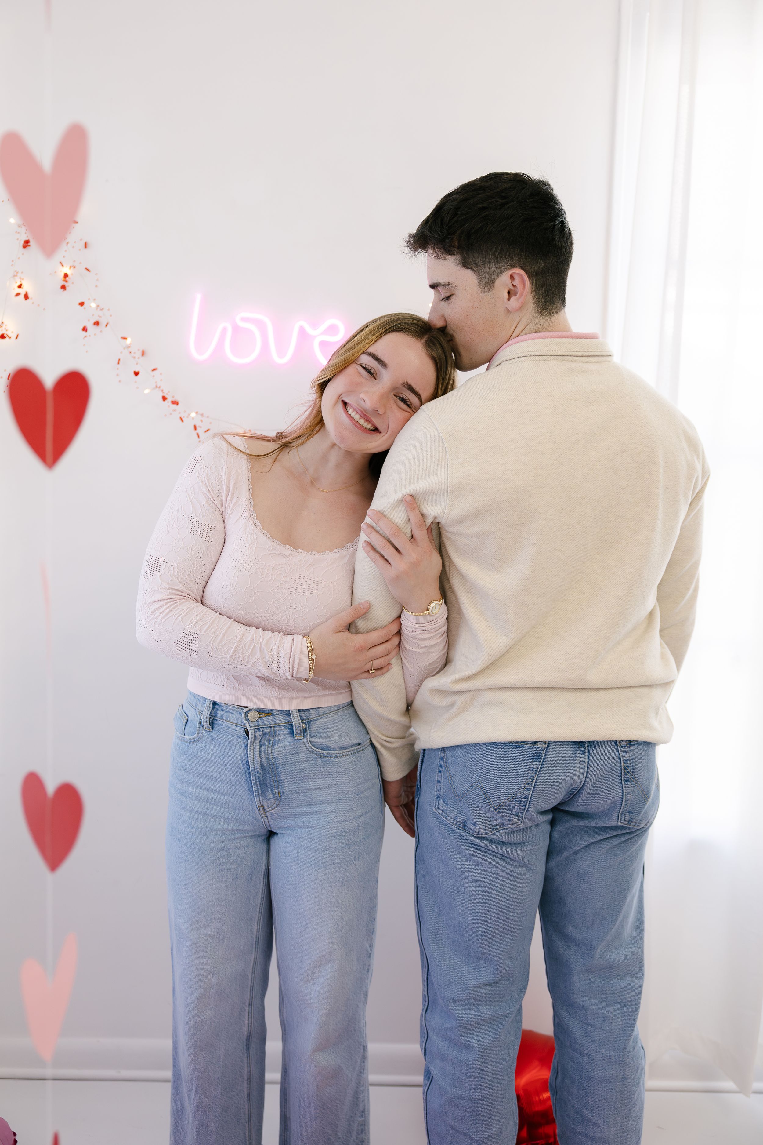 A romantic couple in matching jeans and light tops embrace and laugh together against a wall decorated with heart shapes.