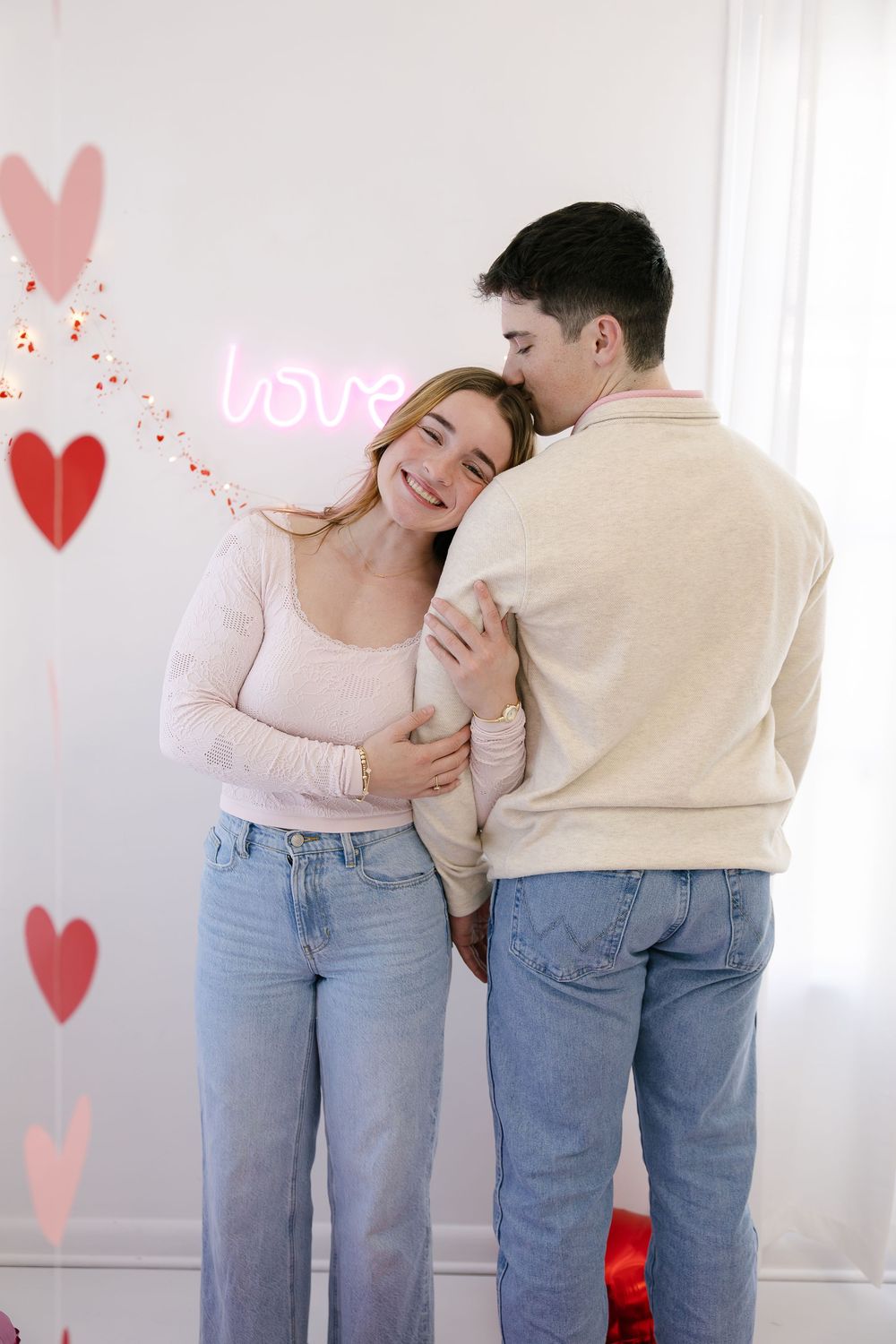 A romantic couple in matching jeans and light tops embrace and laugh together against a wall decorated with heart shapes.