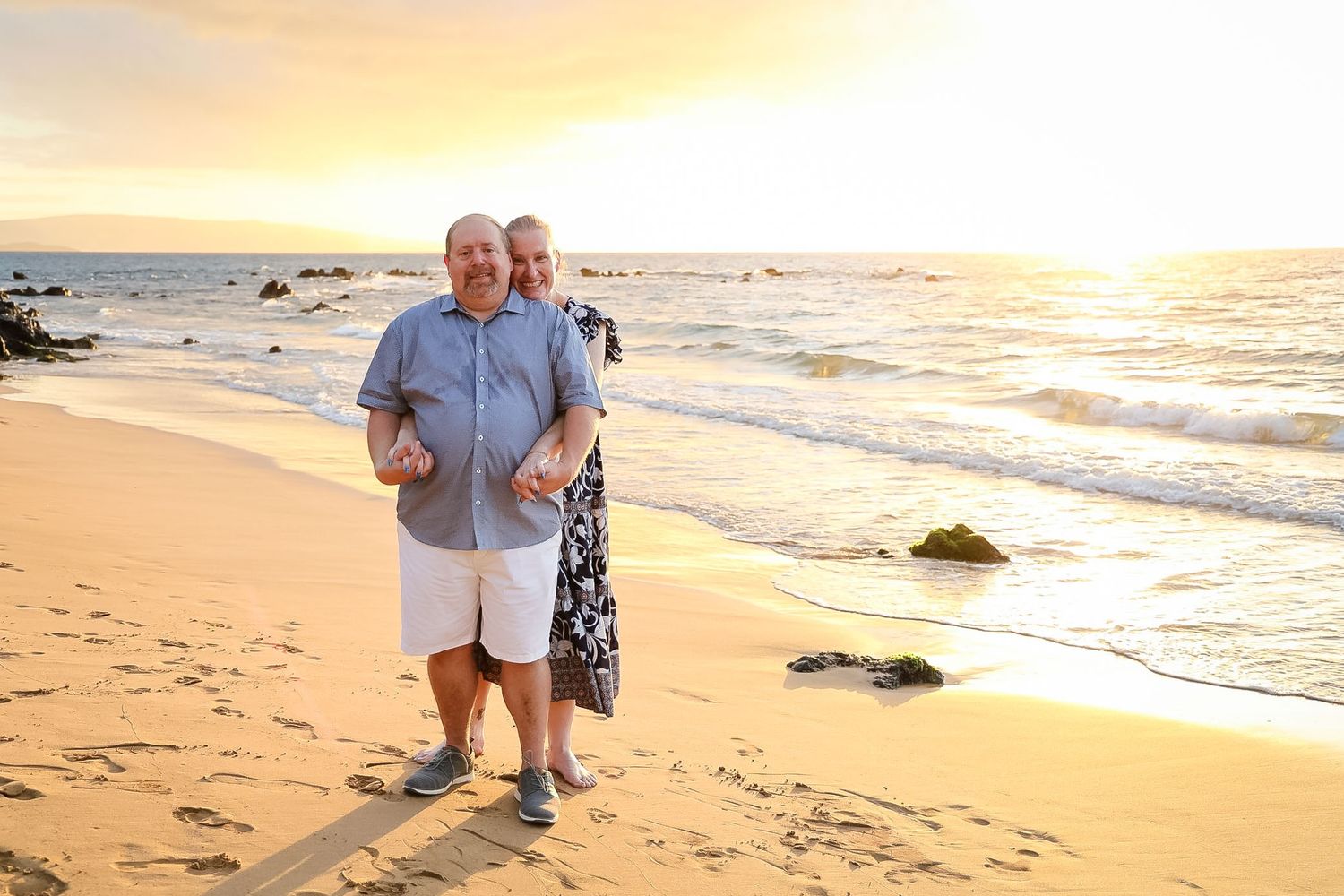 Two people share a tender moment with a baby on a scenic beach at sunset with waves crashing on rocky shore.