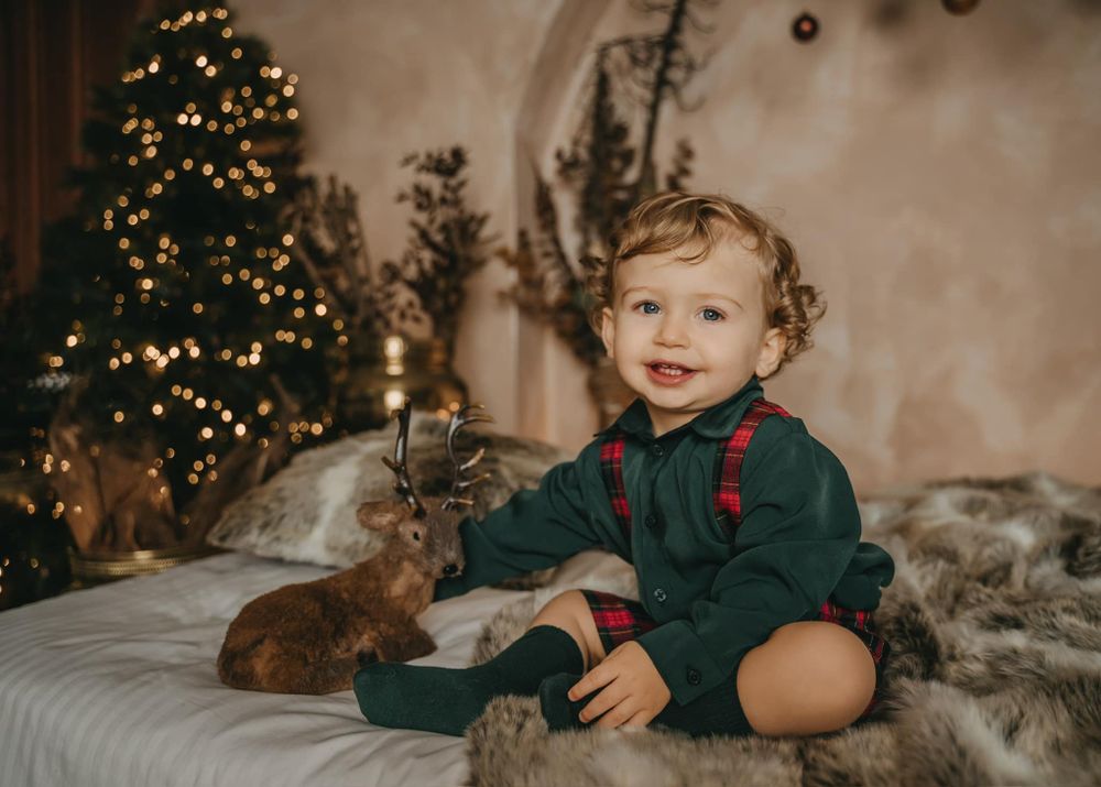 Cozy Christmas scene with toddler in green outfit sitting with bunny on fur blanket near decorated tree.