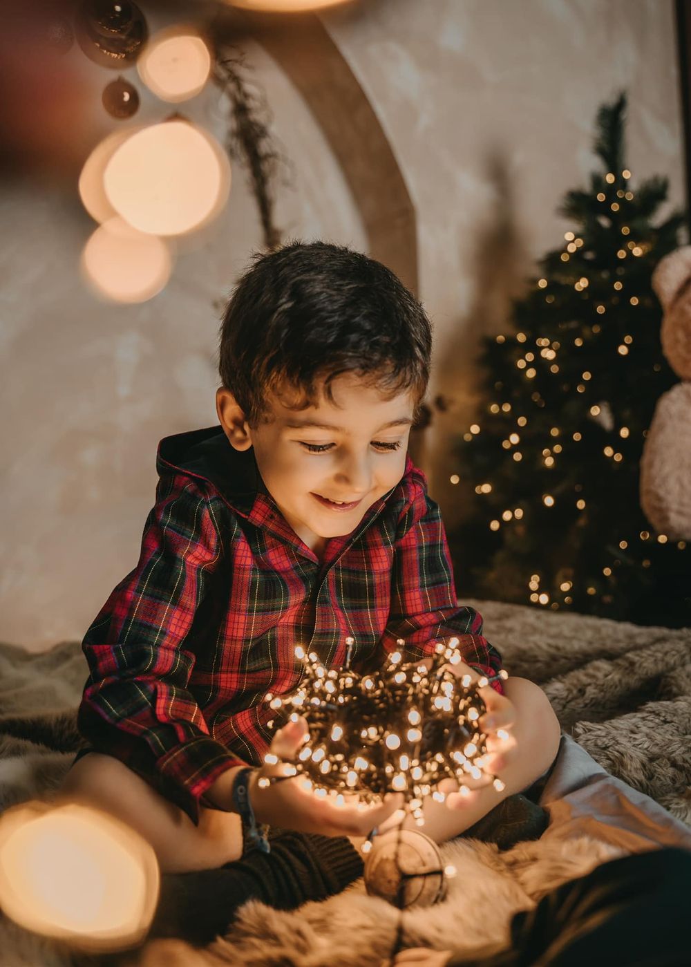 Child in plaid shirt holding glowing fairy lights near Christmas tree.