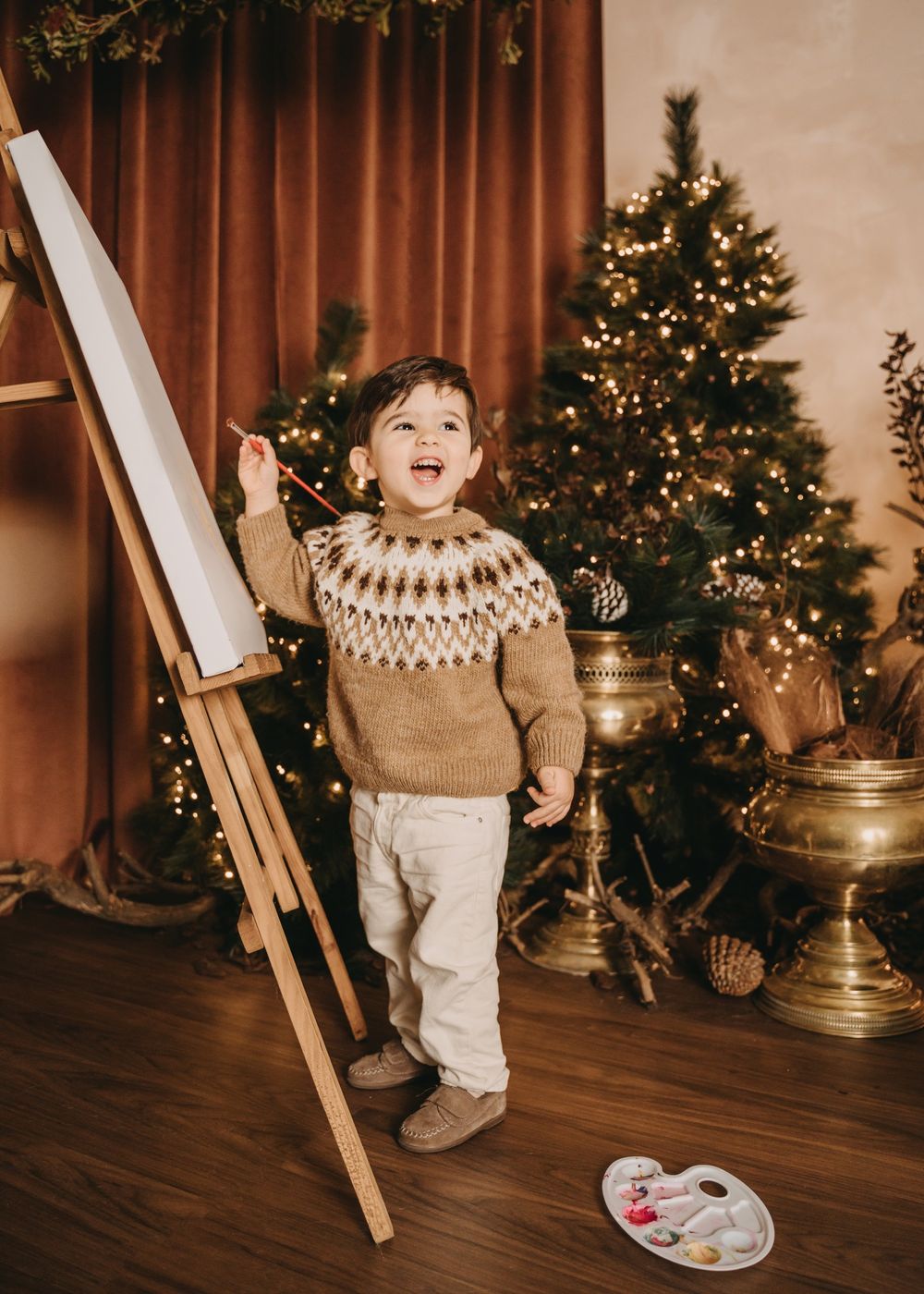 A festive holiday scene with an art easel, Christmas tree, and decorative copper vessels in a warm-toned room.