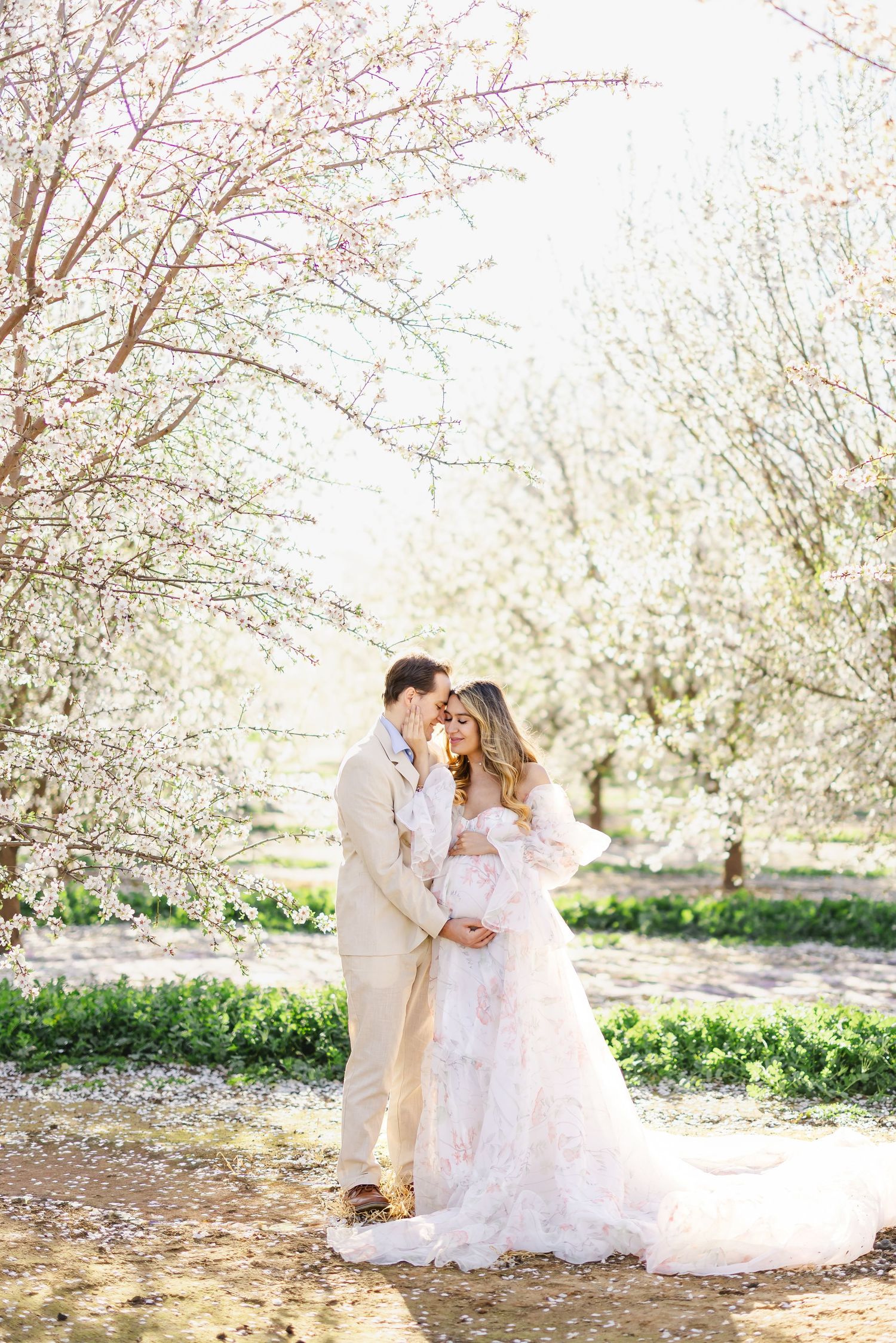 Romantic spring maternity photo under blooming cherry blossom trees with flowing white lace dress and beige suit.