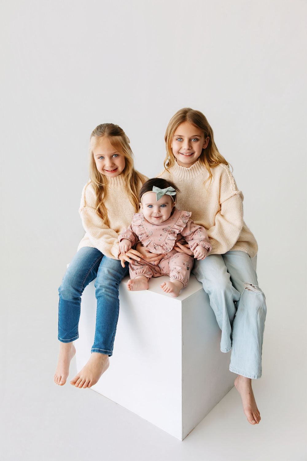 Three siblings in casual clothing sit together on white blocks against a light background.