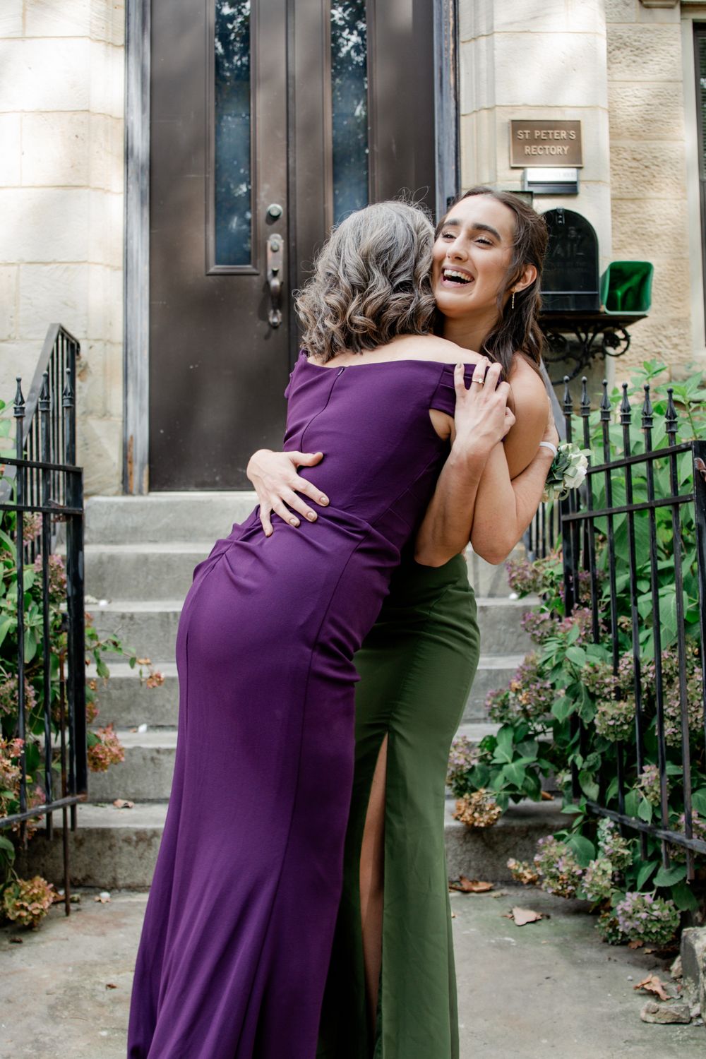 Two women in elegant dresses share a joyful embrace on stone steps outside a front door with wrought iron railings.