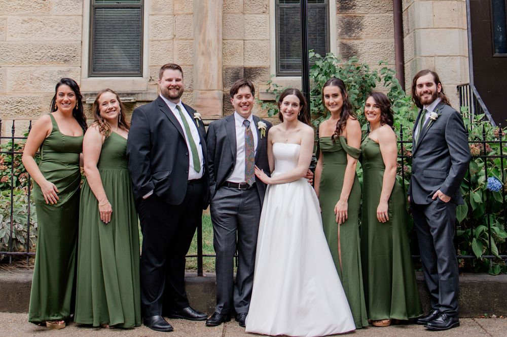 Wedding party poses together outside stone church building wearing olive green dresses and dark suits.