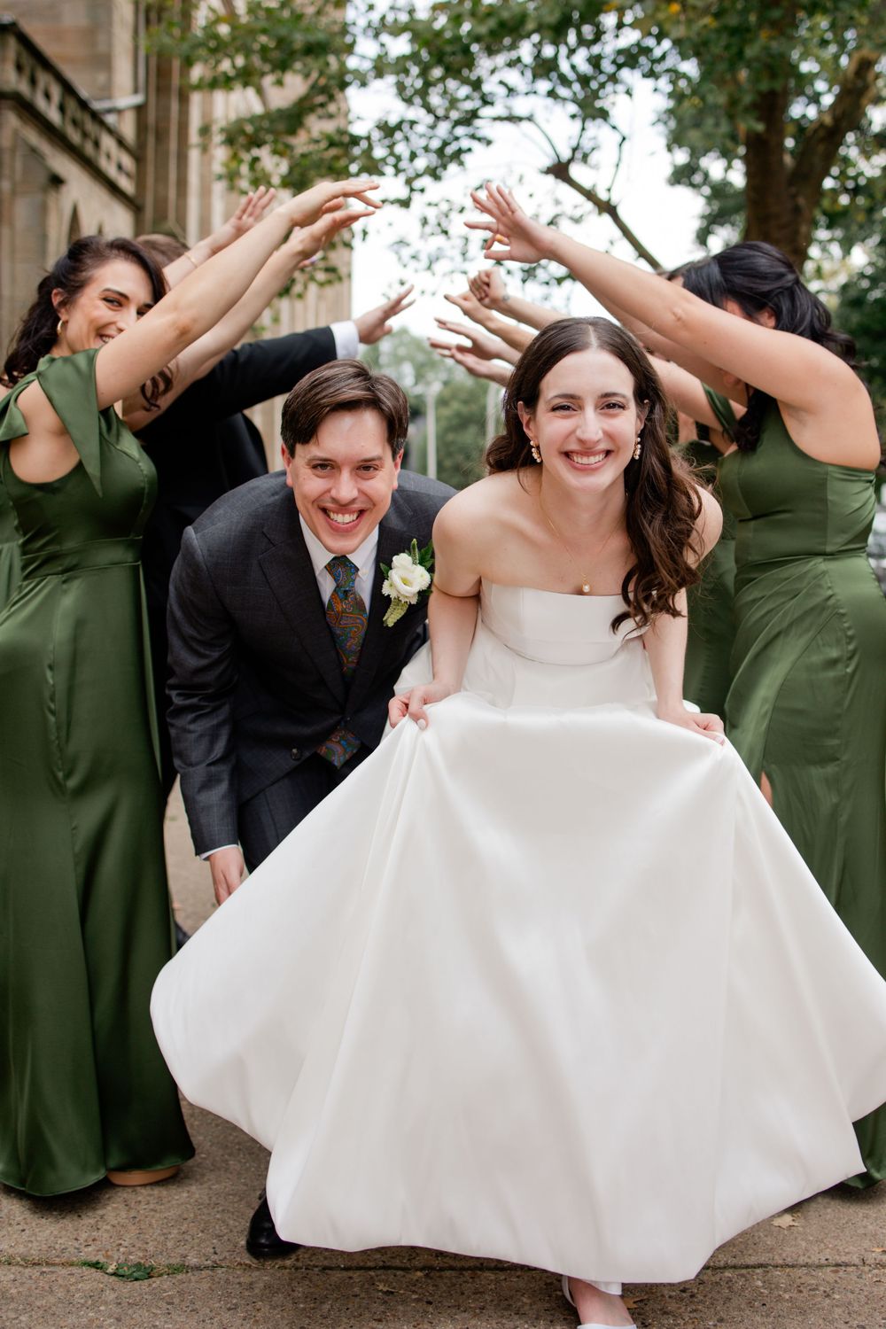 Wedding party in emerald green dresses and formal attire pose playfully on stone steps outdoors.