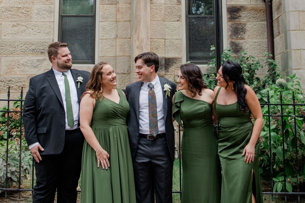 Wedding party in coordinated green dresses and black suits pose together against stone building exterior.