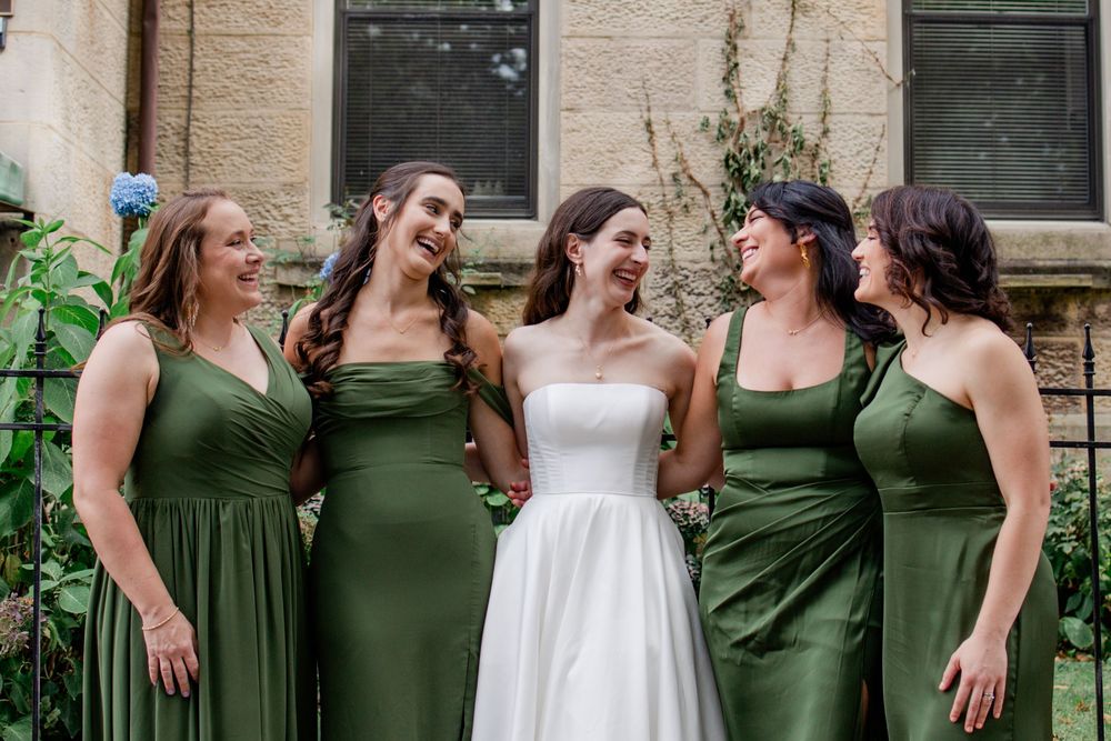 A group of bridesmaids in olive green dresses and one in white laugh together during a wedding photo session.