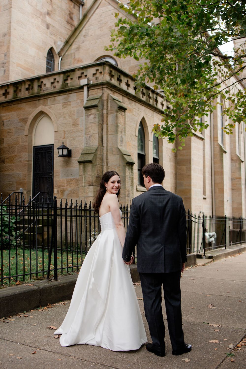 A couple in wedding attire walks along a historic stone church exterior with Gothic architecture details.