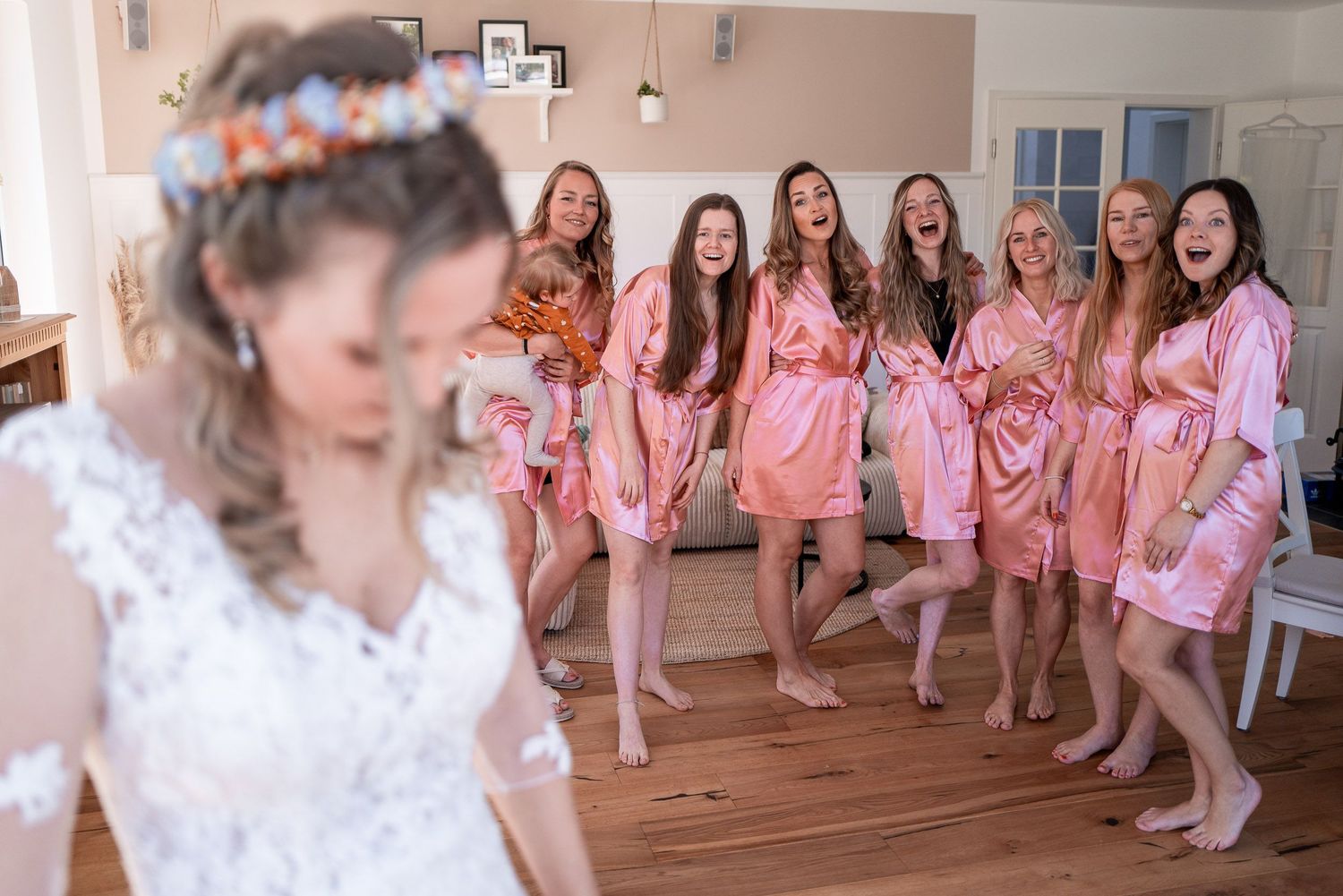 Braut in weißem Spitzenkleid im Vordergrund, dahinter sechs Brautjungfern in rosa Seiden-Morgenmänteln, die gemeinsam lachen und feiern.