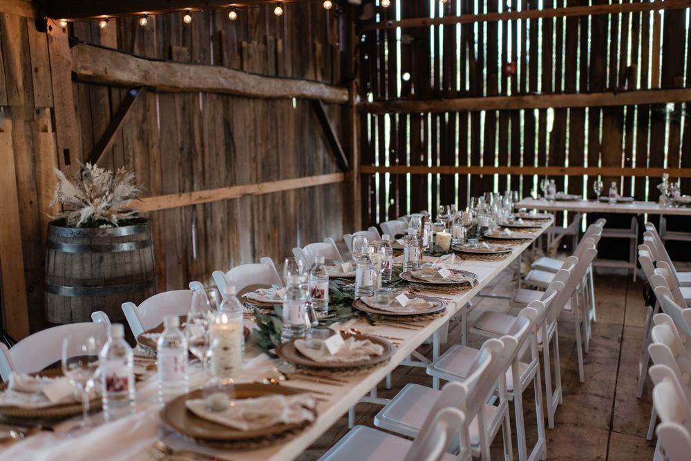 Long rustic wooden tables set for an elegant wedding reception with white linens and natural decor in a barn setting.
