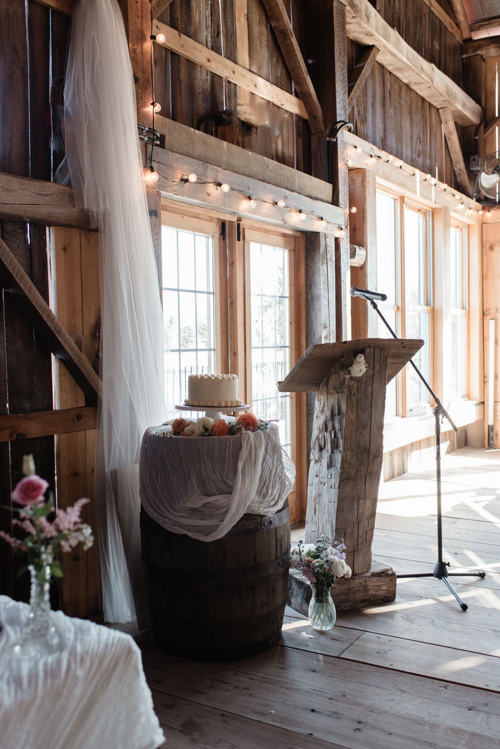 Rustic barn interior with white drapes, wooden beams, and natural light streaming through multiple windows.