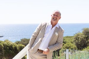 groom wearing a beige suit and white shirt poses outdoors against an ocean backdrop on a sunny day.