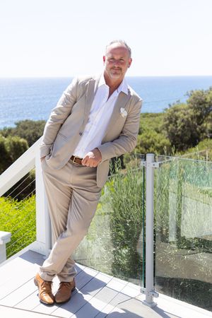 Elegant groom in beige suit posing against ocean view backdrop on sunny terrace.
