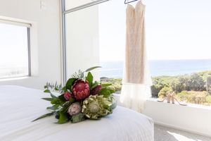A pink and white bridal bouquet rests on a bed near a hanging wedding dress with an ocean view through the window.