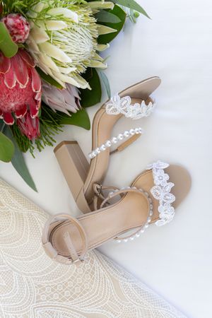 Bridal shoes with delicate lace trim and pearl details next to pink protea bouquet.