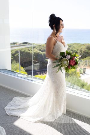 A bride in a flowing lace wedding gown holds a bouquet while standing near large windows with scenic outdoor views.