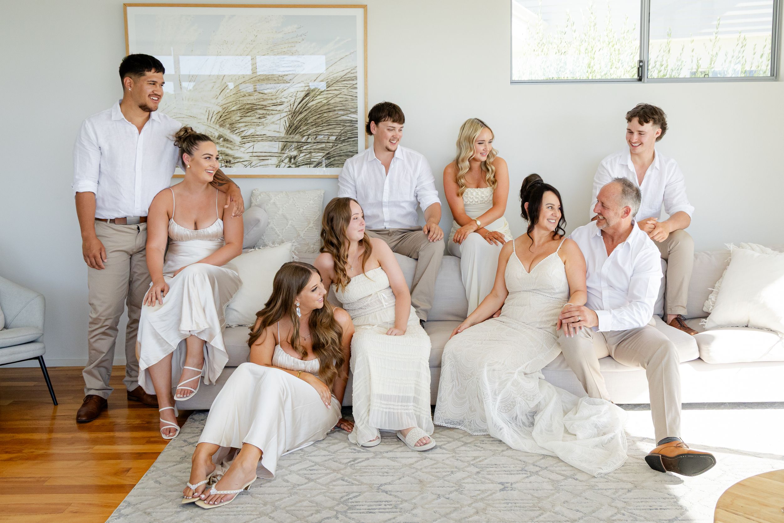 Wedding party dressed in white and beige sits together on a white couch in a bright modern room.