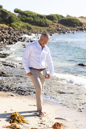 groom in white shirt and khaki pants walking along rocky beach coastline on sunny day.