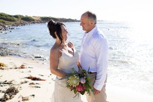 Newlyweds share intimate moments on a scenic beach with waves and rocky coastline in background.