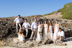 Wedding party in white and beige attire poses together on rocky coastal cliffs near the ocean.