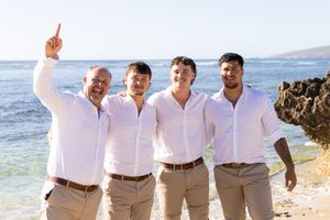 groomsmen in white shirts and khaki pants pose together on a sunny beach with ocean waves and rocks in the background.