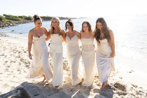 bridemaids in flowing white dresses poses together on sandy beach with ocean view.