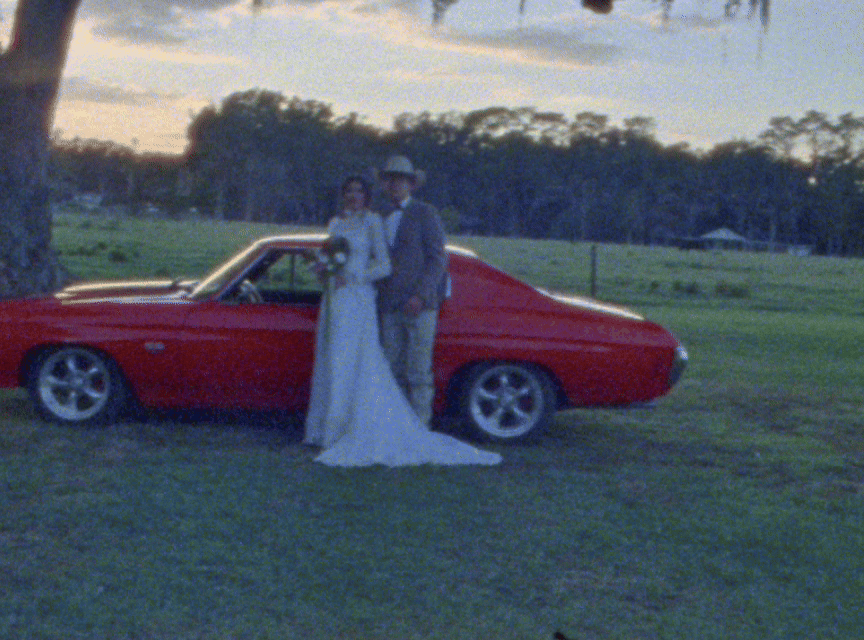 A red classic car parked on grass during sunset with trees in the background.