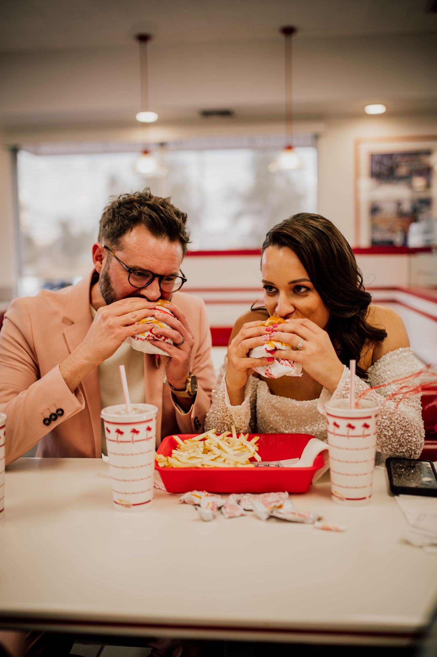 A couple poses playfully outside an In-N-Out Burger restaurant in matching pink outfits during sunset.