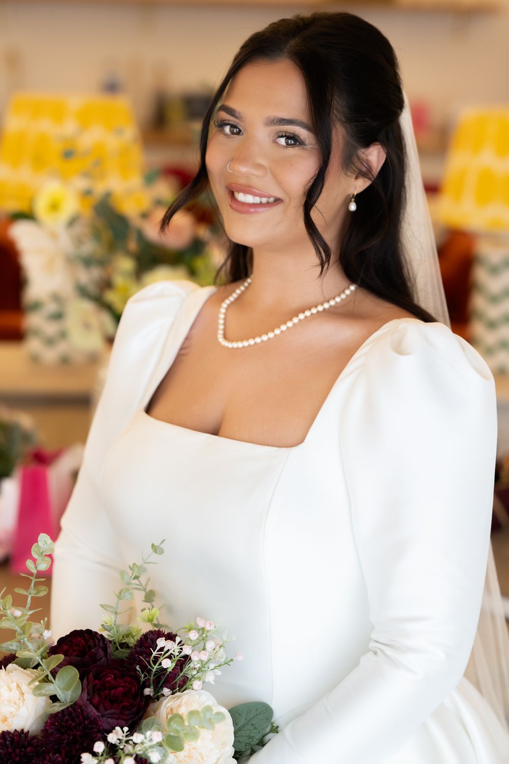 A bride in a classic white long-sleeved wedding dress with pearl necklace holds a deep burgundy and greenery bouquet.