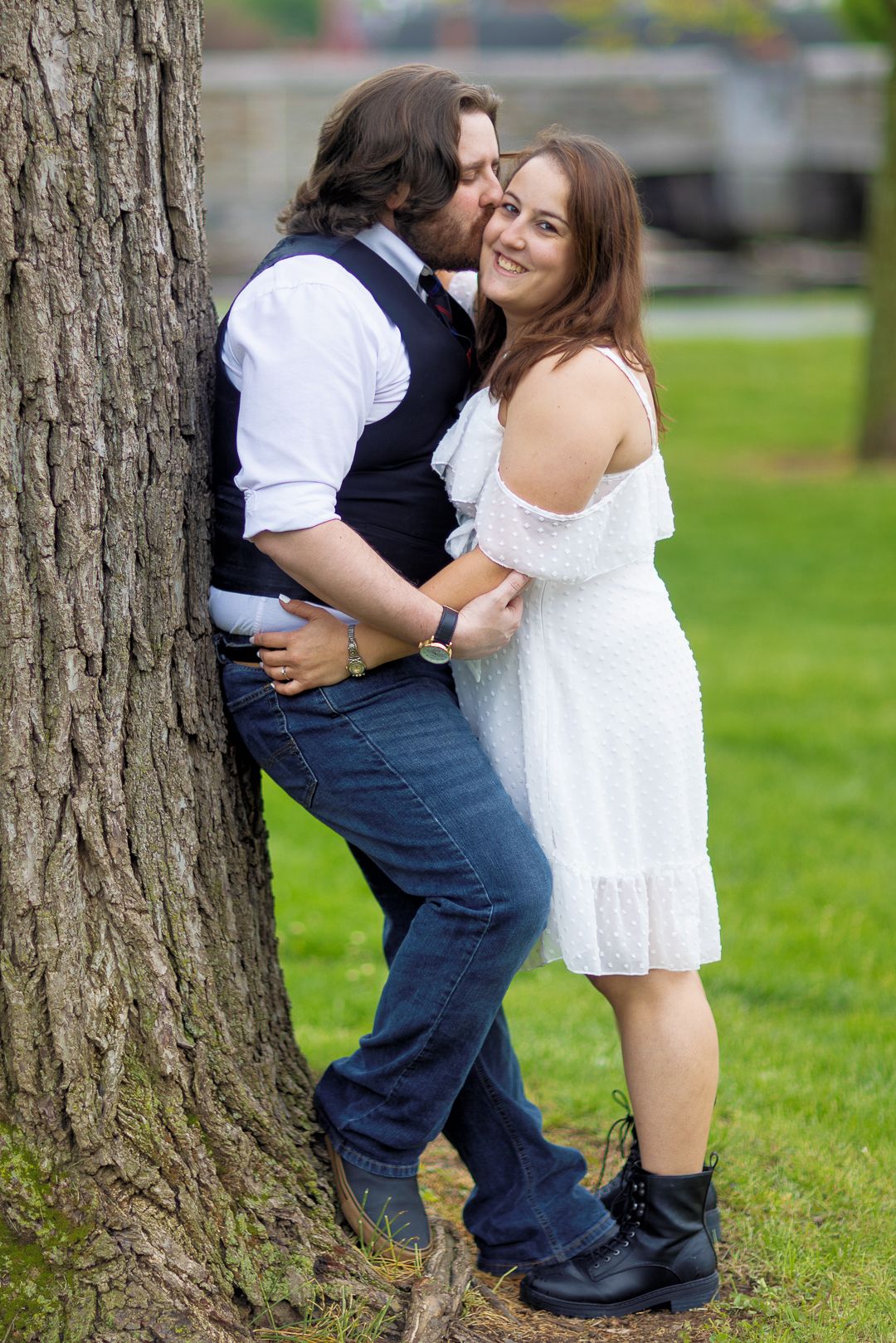 A couple shares tender moments in a series of romantic outdoor photos by a large tree trunk in a grassy park.