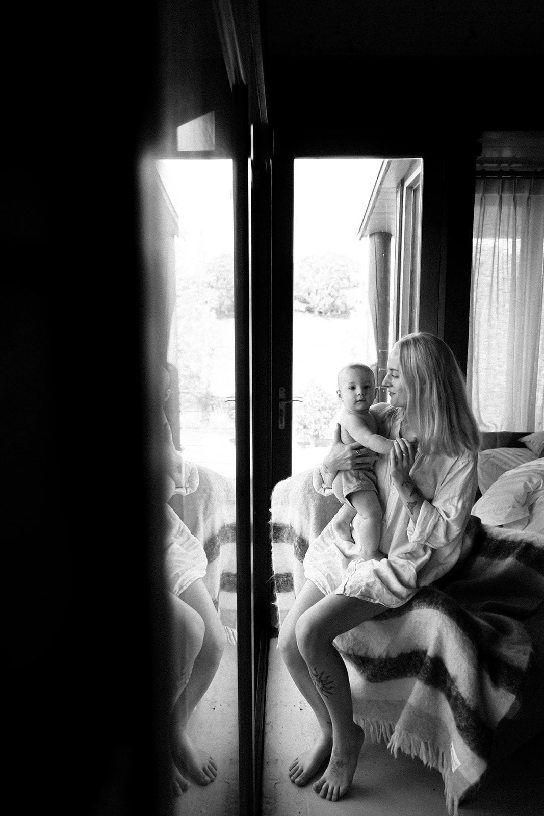 A black and white photo of someone sitting on a bed near a window with natural light streaming in.
