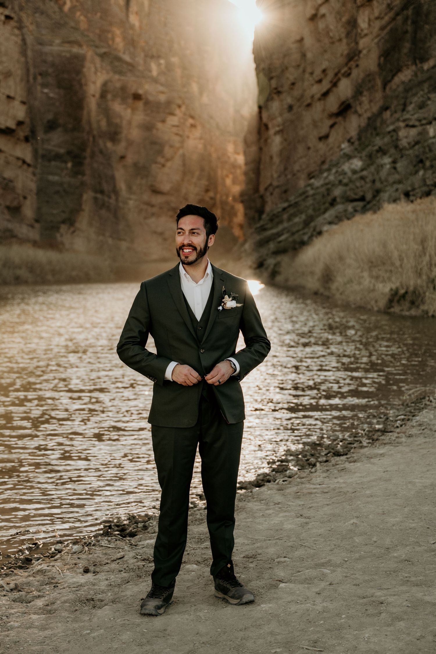 Groom poses confidently on beach with dramatic canyon scenery in background.