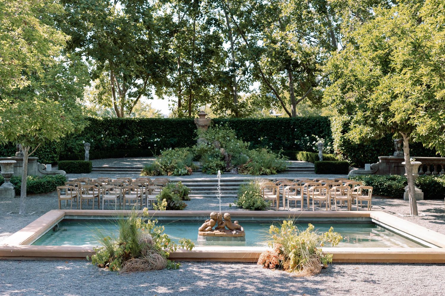 Modern outdoor pool area with stone terrace and fountains surrounded by lush greenery.