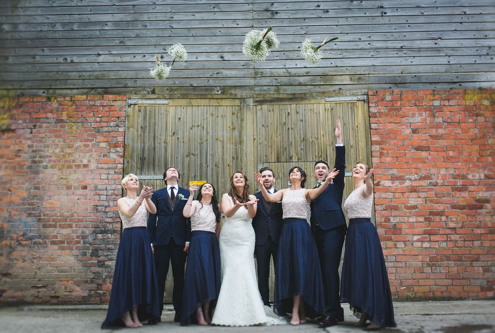Wedding party in navy blue dresses throwing bouquets in the air against a rustic brick wall.
