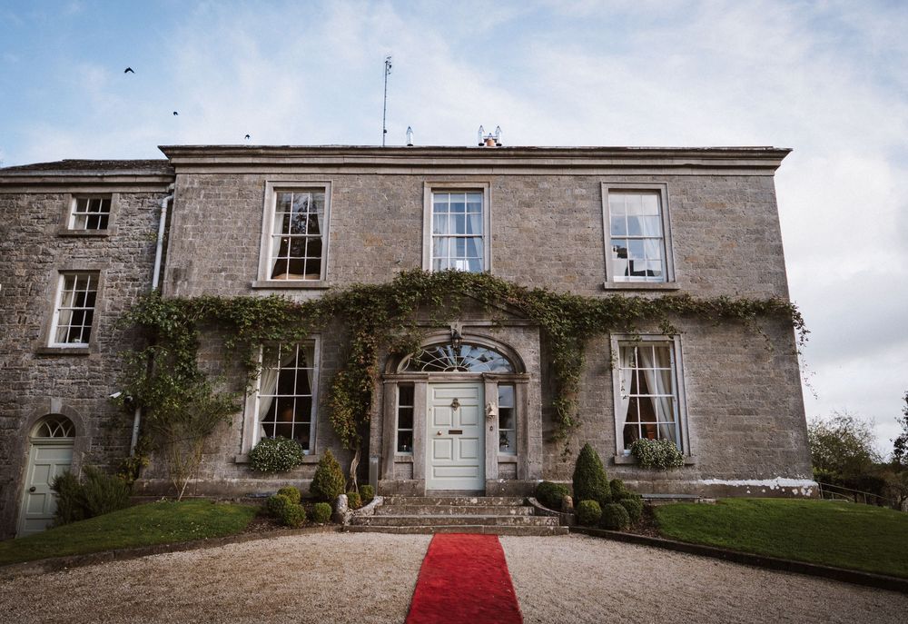 Elegant Georgian manor house with climbing ivy and red carpet entrance leading to white door under cloudy sky.