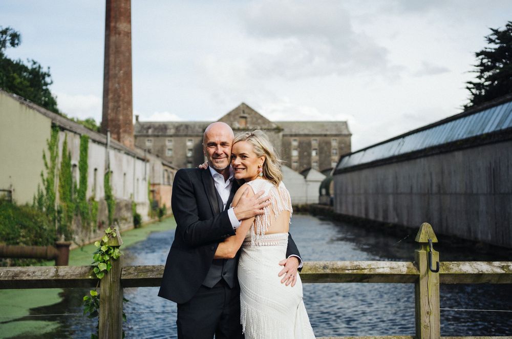 Wedding couple poses along a waterfront with industrial buildings and docks in the background.