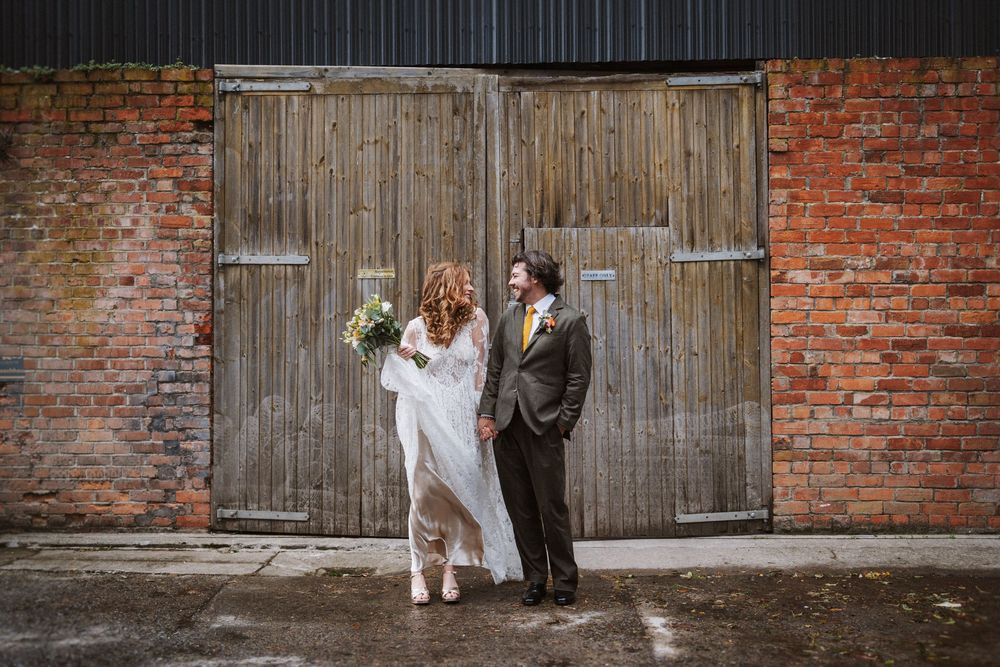 A couple in wedding attire poses together in front of rustic wooden barn doors and brick wall.