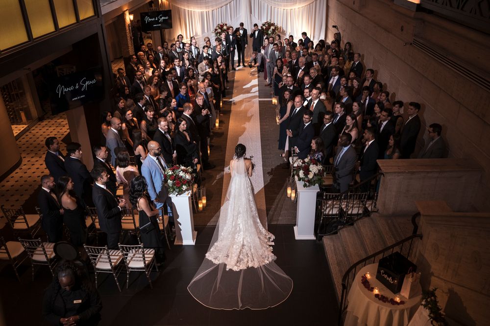A bride in a white gown walks down the aisle during a wedding ceremony at a Union Trust wedding while guests watch from their seats.