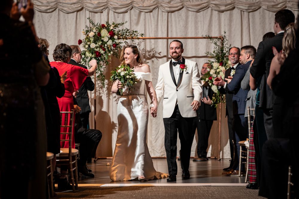 A couple walks down the aisle at their wedding ceremony at Union Trust with elegant floral arrangements and white drapery in the background.