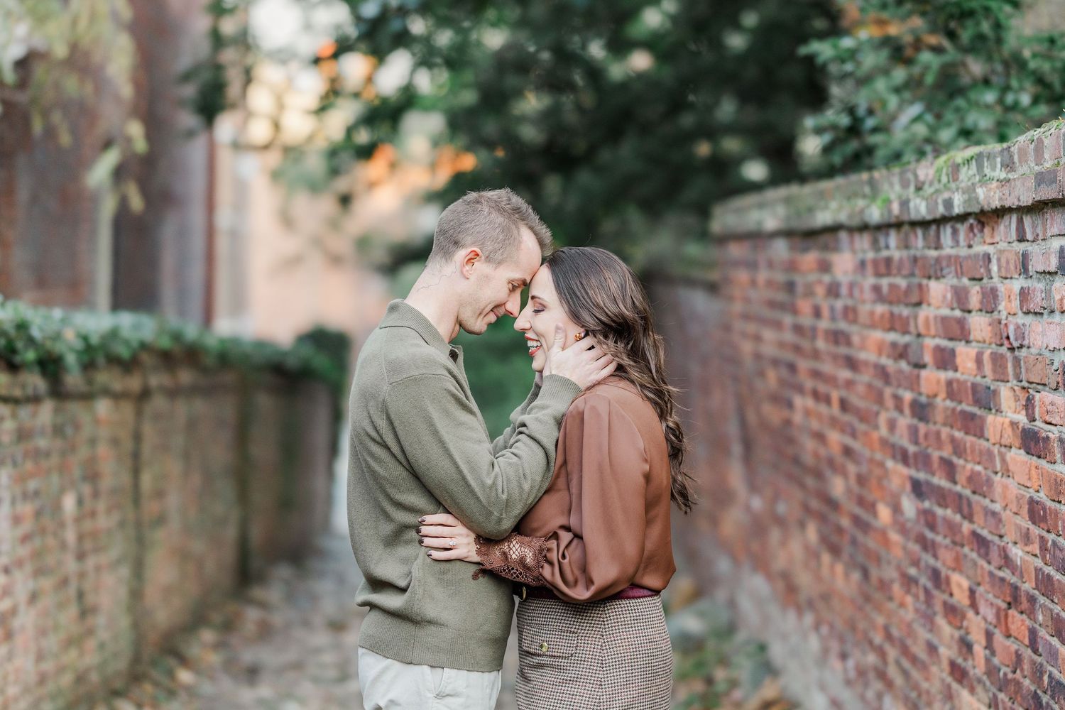A couple shares an intimate moment along a brick-walled path during an autumn engagement photo session.
