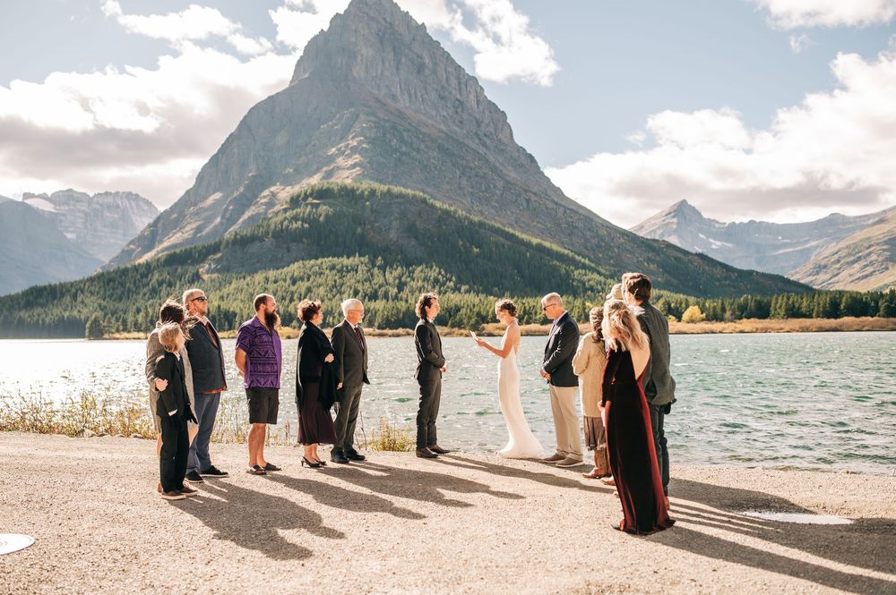 Wedding party stands in line against scenic mountain backdrop at lakeside beach during outdoor ceremony.