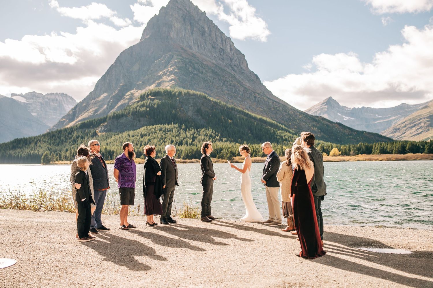 Wedding party stands in line against scenic mountain backdrop at lakeside beach during outdoor ceremony.