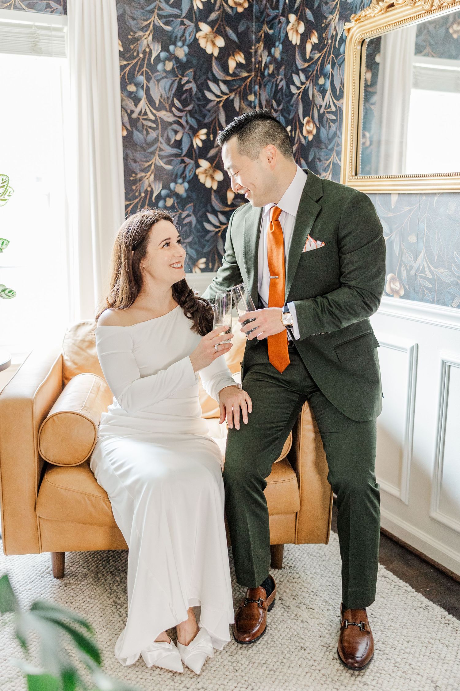 A couple in wedding attire shares a joyful moment while sitting on a tan leather couch in an elegant room.